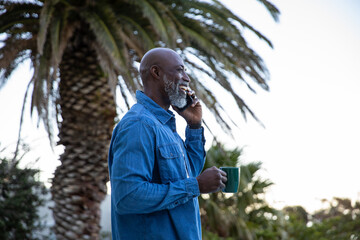 Senior african american man standing in garden by palm fronds, calling on smartphone, green mug © wavebreak3