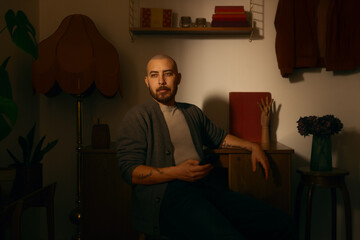 A thoughtful man with tattoos and a beard sits in a dimly lit, vintage-style room using his smartphone. Moody interior with warm lighting and a nostalgic atmosphere.