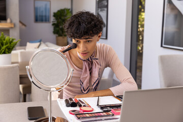 Middle eastern man applying makeup using brush and mirror, checking laptop phone on table at home