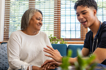 Senior woman and caregiver holding hands on blue sofa at home with stethoscope, plant, water glas
