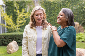 Diverse female friends standing on in backyard chatting and showing wristwatch, hoop earrings