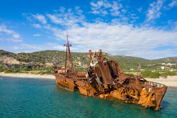 Fototapeta premium The Dimitrios shipwreck lies heavily rusted and broken in shallow turquoise water near a sandy beach, backed by green hills and scattered houses under a bright blue sky in Gythio, Greece.