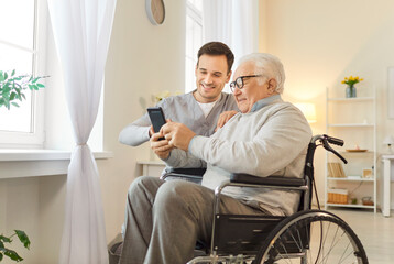 Positive male nurse assisting an elderly patient in a wheelchair with a smartphone and online...