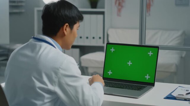 Doctor working on laptop with green screen in medical office.