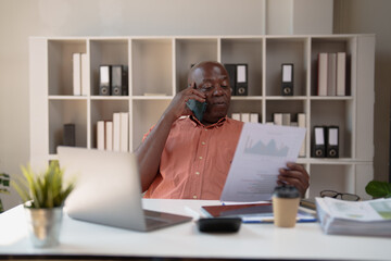 Senior Black businessman in office on a phone call while reviewing financial documents and charts, focused on managing operations, strategy and data-driven decisions at his desk