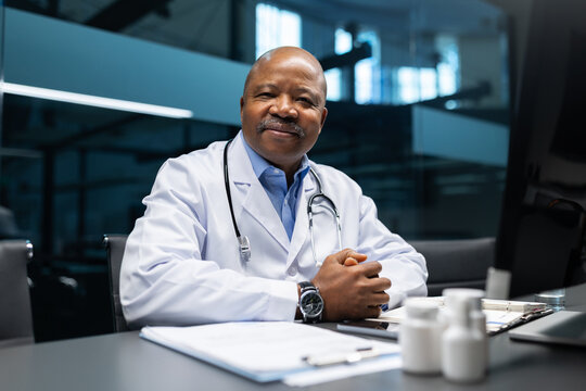 Black mature doctor sitting confidently at office desk wearing lab coat and stethoscope, professional portrait showing leadership, trust, and medical business lifestyle