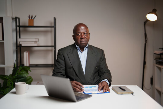 Experienced African American businessman focused on financial data, analyzing charts and graphs on a clipboard while sitting at a white desk with a laptop and coffee mug in a modern office - Powered by Adobe