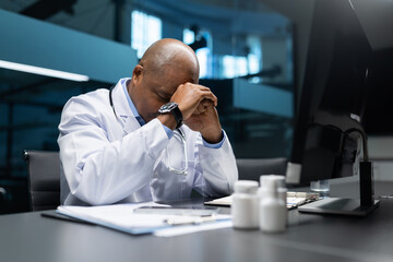 Stressed black mature doctor in lab coat resting head on hands at office desk, showing exhaustion and pressure in professional healthcare business environment