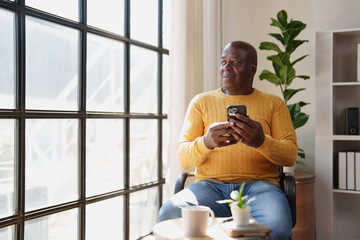 Senior black man relaxing at home, holding a smartphone while looking pensively out of a large window, enjoying a peaceful moment of contemplation indoors