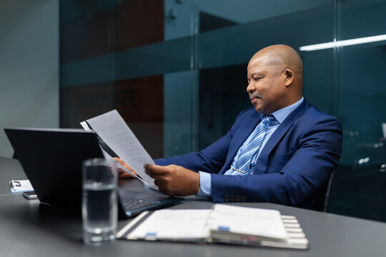 Black mature businessman reading paperwork while leaning back in office chair, calm and confident executive evaluating information in corporate environment