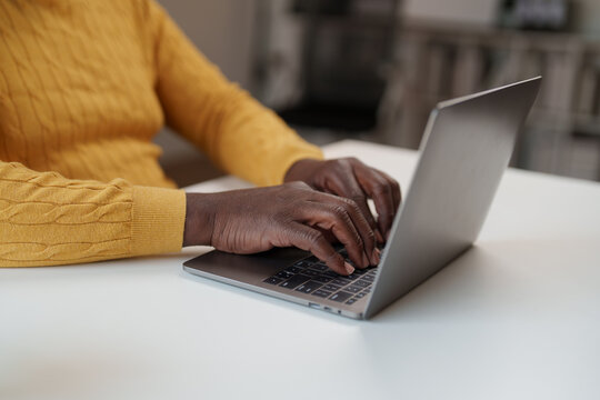 Dark-skinned hands typing on a laptop keyboard at a white desk, remote work or study in a cozy home office with a yellow sweater and connected digital lifestyle - Powered by Adobe