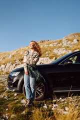 Traveler in hat stands next to black car in a mountainous area.  Beautiful woman on road trip feels...