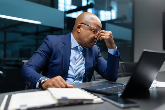 Tired black mature businessman rubbing his eyes at desk while working on laptop, illustrating exhaustion, stress, and high responsibility in executive corporate work