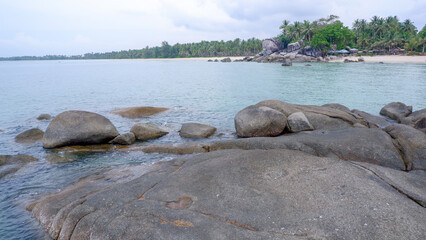 A beautiful coastal landscape featuring granite rock formations in the foreground and a dense coconut palm forest along Siangau Beach