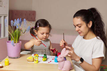 Happy mother and cute little child girl are preparing to celebrate Easter at home. Mom and daughter painting eggs before Easter together.