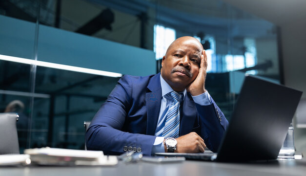 Thoughtful black mature businessman resting his head on his hand while working at a laptop in a glass office, expressing focus, decision making, and leadership in a corporate environment