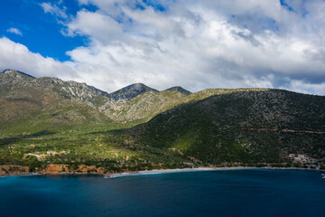 Fototapeta premium A vibrant blue bay borders a secluded stretch of coastline, backed by lush, sun-dappled mountains under a sky of dramatic clouds. The landscape features rugged slopes, scattered houses, and a tranquil