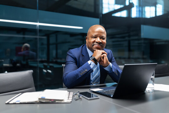 Confident black mature businessman sitting at conference table with hands folded, looking at camera in modern office, representing leadership, professionalism, and corporate success