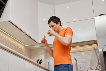 Young man standing in a modern kitchen, eating a sandwich and drinking milk