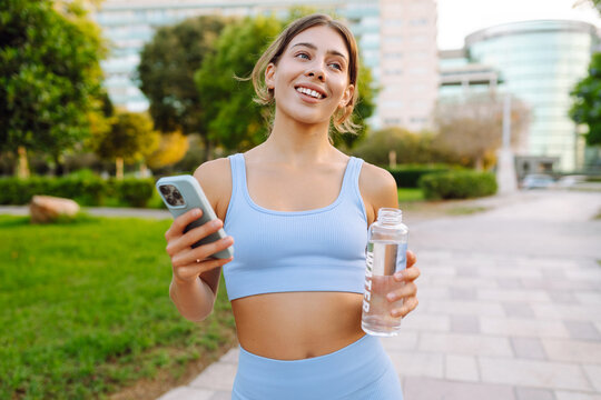 Cheerful woman in sportswear stands in a sunny park with a phone and a bottle of water in hands. Beautiful woman drinks water and texts after workout. Concept: sports, recreation. Active lifestyle.