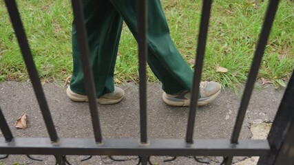 green pants walking behind fence along cracked sidewalk, worn sneakers scuffing pavement, scattered autumn leaves on grass, vertical iron bars creating framed barrier, low angle candid crop of legs