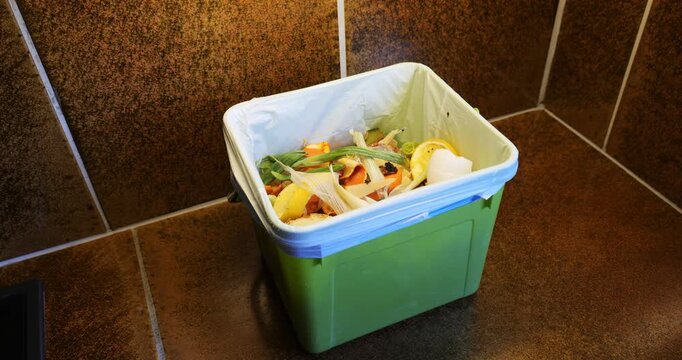 Cooking food, peeled vegetable leftovers in a plastic trash container inside domestic kitchen counter top. Food recycling concept, composting, biofuel, home waste sorting, real time pan shot