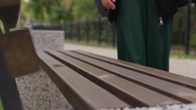 park bench closeup person approaching, candid sequence shows individual walking toward wooden bench on gravel path, hand reaching for seat edge, wearing green trousers and dark coat, iron fence