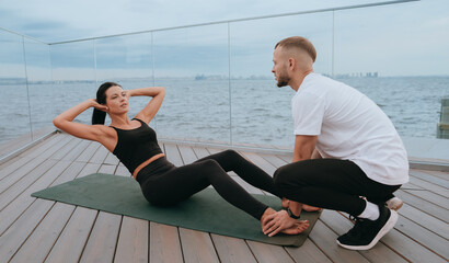 Man provides support during a partner's exercise on a deck overlooking the ocean, highlighting teamwork and fitness motivation.