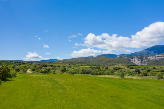 Wide green fields stretch toward rugged mountains and rolling hills under a vivid blue sky dotted with fluffy white clouds near Bierge, Spain. The open landscape is bathed in bright sunlight, creating