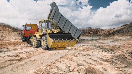 Fototapeta premium Heavy equipment works on a construction site with earthen embankments, against a backdrop of shifting clouds. A dump truck delivers earth, and a loader levels it