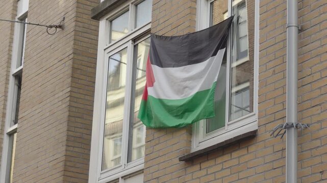 A Palestinian flag is displayed from an upper window of a brick residential building. The peaceful protest gesture is set in a calm urban street in daylight - Amsterdam, De Jordaan