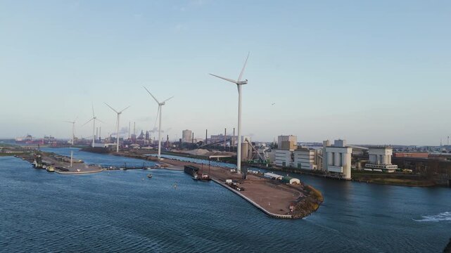 Drone footage showing wind turbines positioned along a waterfront near the Tata Steel industrial complex. The shot includes blue water, coastal infrastructure, and distant steel plant structures in an