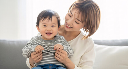 Happy young Asian mother holding her smiling baby son on a comfortable sofa at home, sharing a moment of joy and love. Warm, bright indoor lighting.