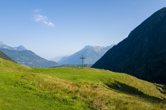 A solitary wooden cross stands atop a sunlit green hill with sweeping views of distant alpine peaks and deep valleys near Champex, Switzerland under a clear blue sky.