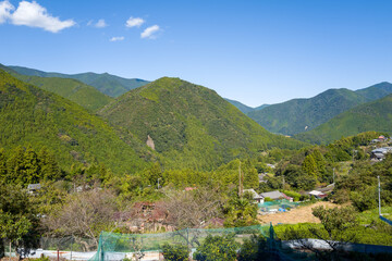A panoramic view of a small rural village nestled among lush green mountains and dense forest under a vivid blue sky along the Kumano Kodo trail in Wakayama, Japan.