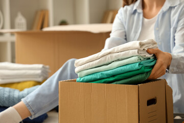 Young woman packing stack of clothes into cardboard box in bedroom, closeup
