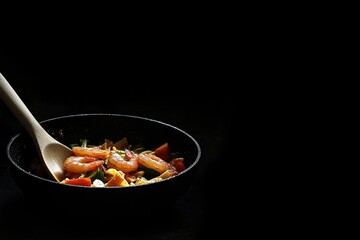 A vibrant dish of stir-fried vegetables and shrimp sizzling inside a black wok with a wooden handle, set against a dark background.