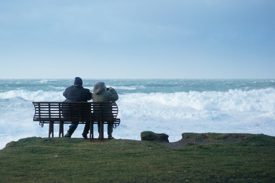 senior couple sitting on bench watching big Atlantic storm waves in Cornwall UK