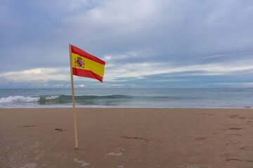 A Spanish flag stands in the sand of a wide, empty beach, waving gently in the breeze. The sea stretches to the horizon under a cloudy, overcast sky, creating a serene coastal scene with subtle colors