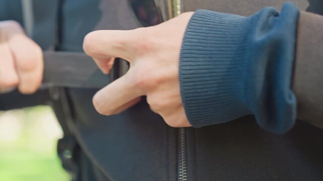 hands fastening backpack chest strap closeup, courier adjusts buckle and zipper on dark jacket, textured nylon webbing pulled tight with thumb and fingertip, sunlight glint on fabric, park background