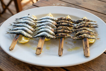 Plato de Espetos de Sardinas Asadas acompañadas de Rodajas de Limón servidas en una Mesa de madera frente a las brasas en la Playa © Iombi