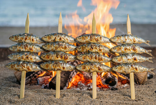 Primer plano de un espeto de sardinas as&aacute;ndose a la brasa con el fuego de una hoguera de fondo en la playa