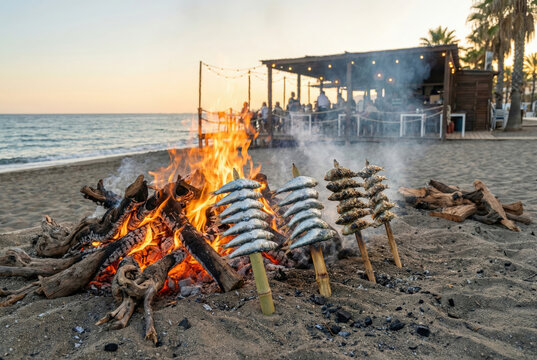 Varias hileras de espetos de sardinas clavados en la arena as&aacute;ndose al calor de una gran hoguera frente a un chiringuito al atardecer