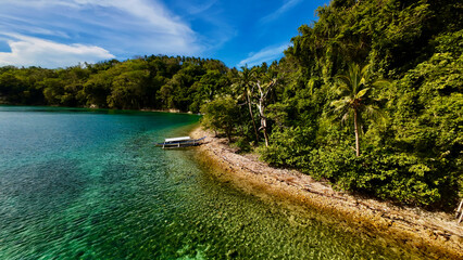 Small boat moored along a tropical shoreline with crystal clear water. Aerial view shows lush green jungle, palm trees, rocky coast, and shallow turquoise sea under blue sky.