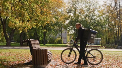 Delivery courier with bicycle in park pauses to secure bike by wooden bench among golden autumn leaves backpack parcel visible soft sunlight filters through trees quiet street and nearby
