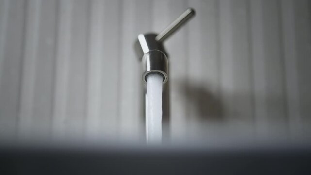 Close-up of minimalist metal faucet in modern bathroom. Human hand turns on and off tap and steady stream of clear water flows into sink. Soft light, gray textured wall, clean interior details