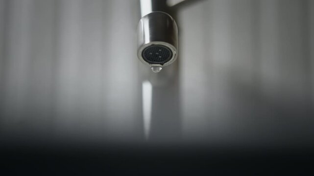 Close-up bathroom faucet with water slowly dripping drop by drop. Steady drip emphasizes water loss caused by an improperly closed or faulty tap. Focus on falling droplets, water waste, mindful usage