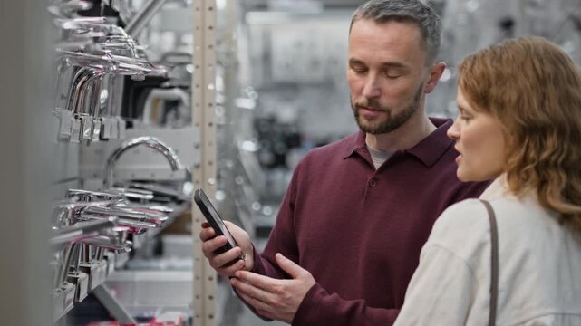 Side view pf Caucasian wife and husband looking for silver tap while comparing all available taps with photo on smartphone