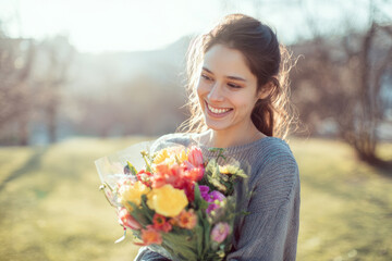Woman smiling while holding bouquet of spring flowers in bright outdoor park, soft sunlight, casual clothing, joyful International Women's Day lifestyle photography