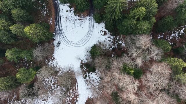 Top-down drone flight over a mountain road hairpin curve with patchy snow, revealing parallel uphill and downhill roads and dense evergreen forest in a winter alpine landscape.